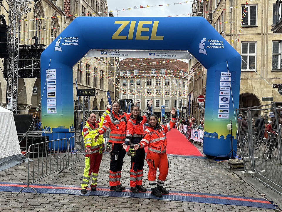 Vier Einsatzkräfte in Einsatzkleidung der vier verschiedenen Hilfsorganisationen posieren lachend unter dem Zielbogen. Im Hintergrund säumen Zuschauer und Absperrgitter den roten Teppich vor dem Ziel.