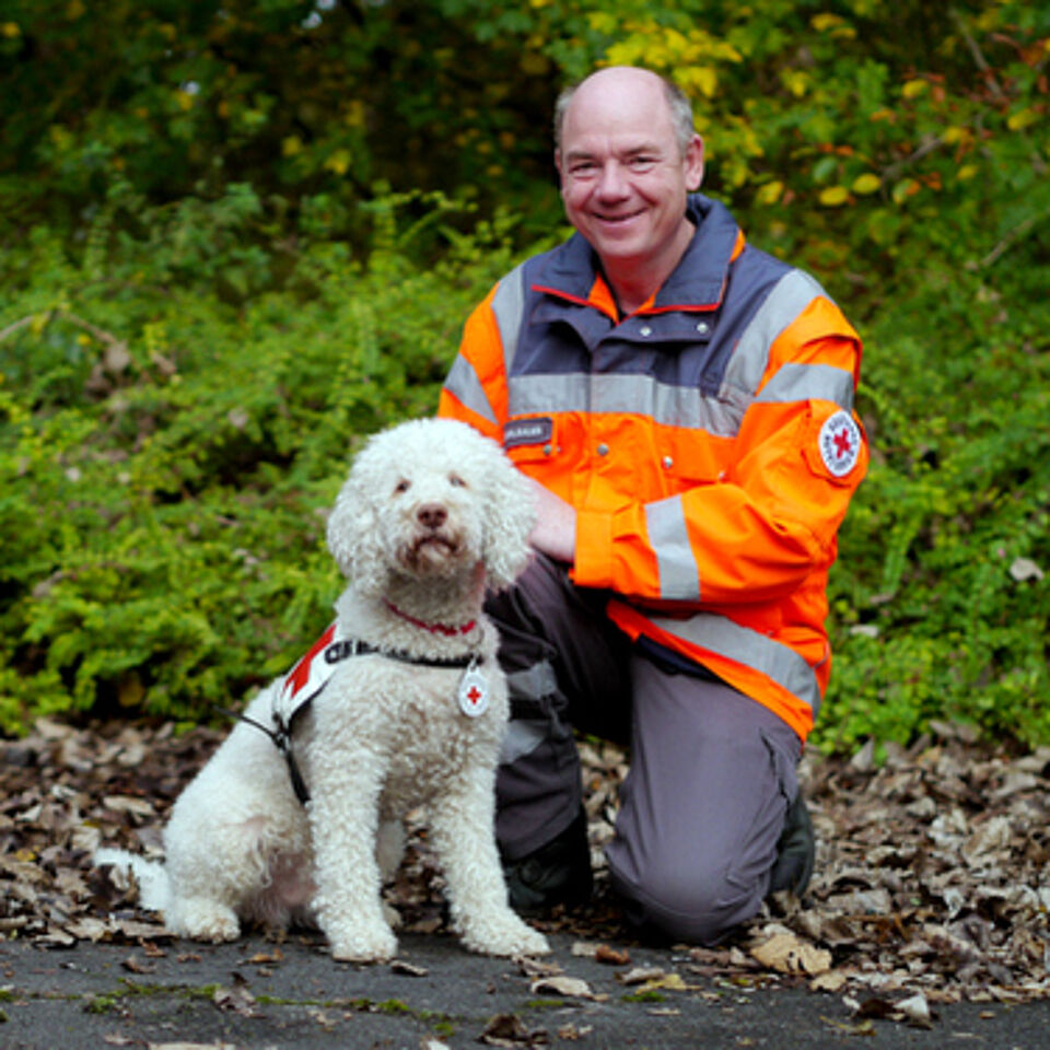 Eine Person in Einsatzkleidung kniet neben einem Hund. Im Hintergrund befindet sich grüne Vegetation.