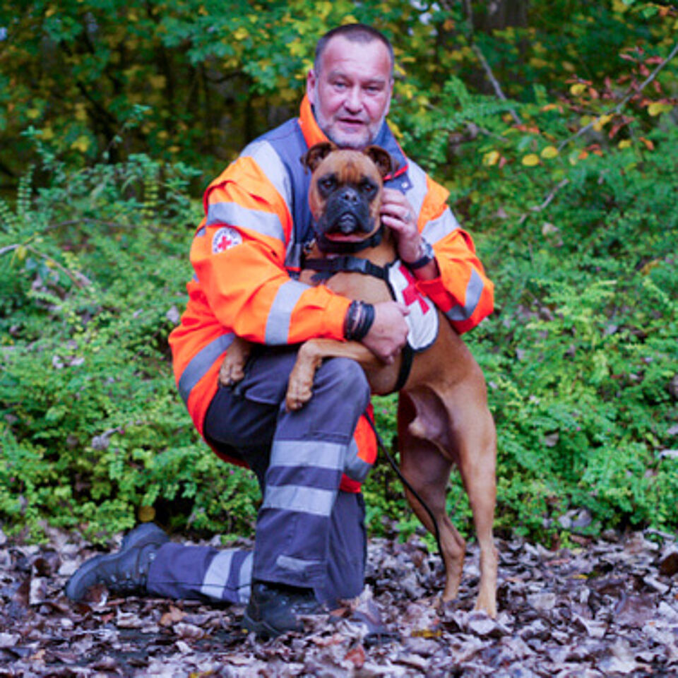 Eine Person in Einsatzkleidung kniet neben einem Hund. Im Hintergrund befindet sich grüne Vegetation.