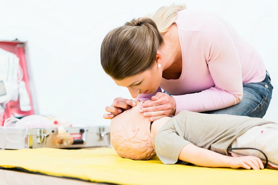 Eine Person führt an einer kindgroßen Übungspuppe eine Wiederbelebung durch. Dabei wird die Nase der Puppe zugehalten und Luft in den Mund geblasen. Im Hintergrund befinden sich medizinische Geräte, darunter ein Defibrillator und weiteres Notfallmaterial
