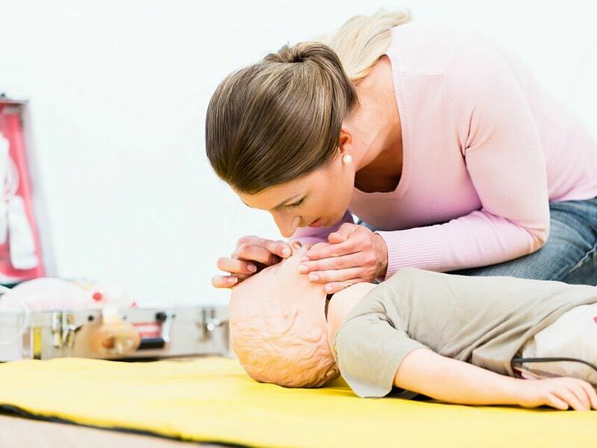 Eine Person führt an einer kindgroßen Übungspuppe eine Wiederbelebung durch. Dabei wird die Nase der Puppe zugehalten und Luft in den Mund geblasen. Im Hintergrund befinden sich medizinische Geräte, darunter ein Defibrillator und weiteres Notfallmaterial