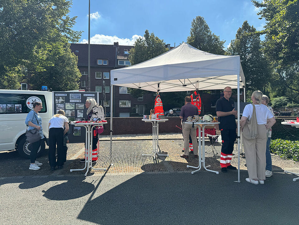 Ein Infostand mit einem Pavillion, einer Stellwand, einem Fahrzeug und mehreren Stehtischen. Mehrere Personen sind miteinander im Gespräch.