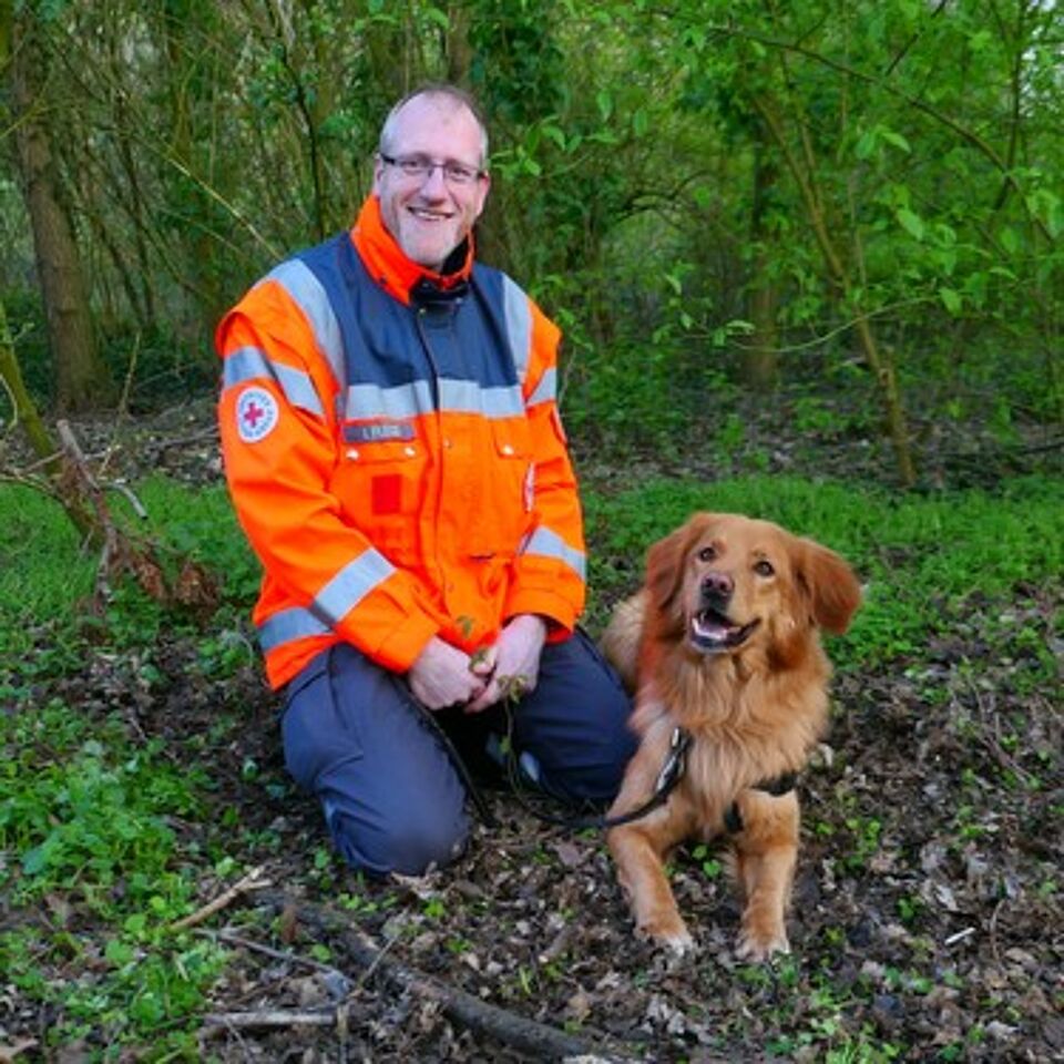 Eine Person in Einsatzkleidung kniet neben einem Hund. Im Hintergrund befindet sich grüne Vegetation.Eine Person in Einsatzkleidung kniet neben einem Hund. Im Hintergrund befindet sich grüne Vegetation.