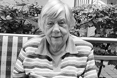 Eine ältere Frau sitzt auf einer Bank, im Hintergrund eine Hecke. Das Bild ist schwarz-weiß.