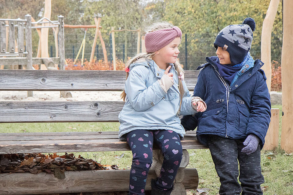 Zwei Kinder in warmer Kleidung sitzen nebeneinander auf einer Holzbank, im Hintergrund ist ein Spielplatz zu sehen