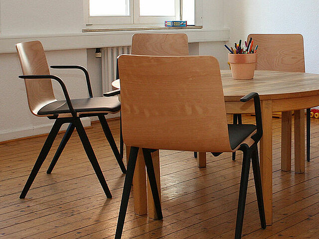 Ein Raum mit einem runden Holztisch, umgeben von sechs Stühlen mit Holzsitzen und schwarzen Metallbeinen. Auf einem Stuhl liegt ein rotes Sitzkissen. In der Mitte des Tisches steht ein Becher mit bunten Stiften. Der Raum hat Holzfußboden, weiße Wände, zwei Fenster mit Tageslicht und einen Heizkörper. Im Hintergrund befinden sich Spielzeug oder Gegenstände an der Wand.