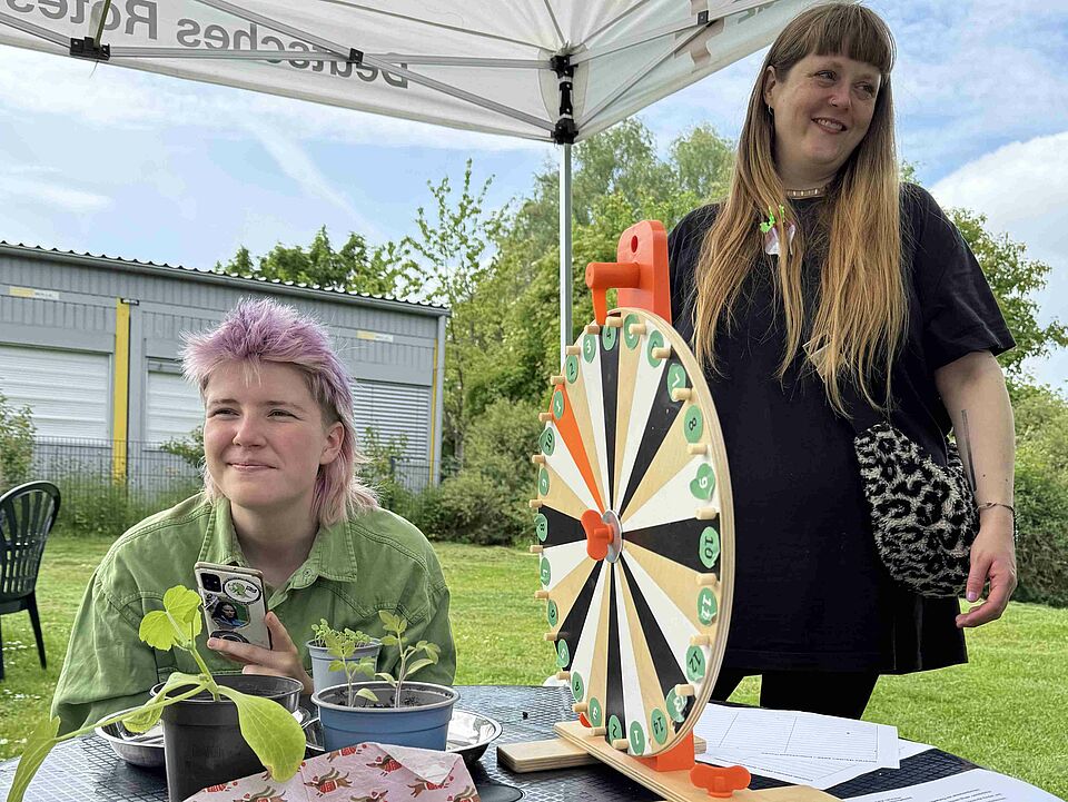 Zwei personen unter einem Pavillion und an einem Tisch, auf dem ein Glücksrad und Pflanzen stehen