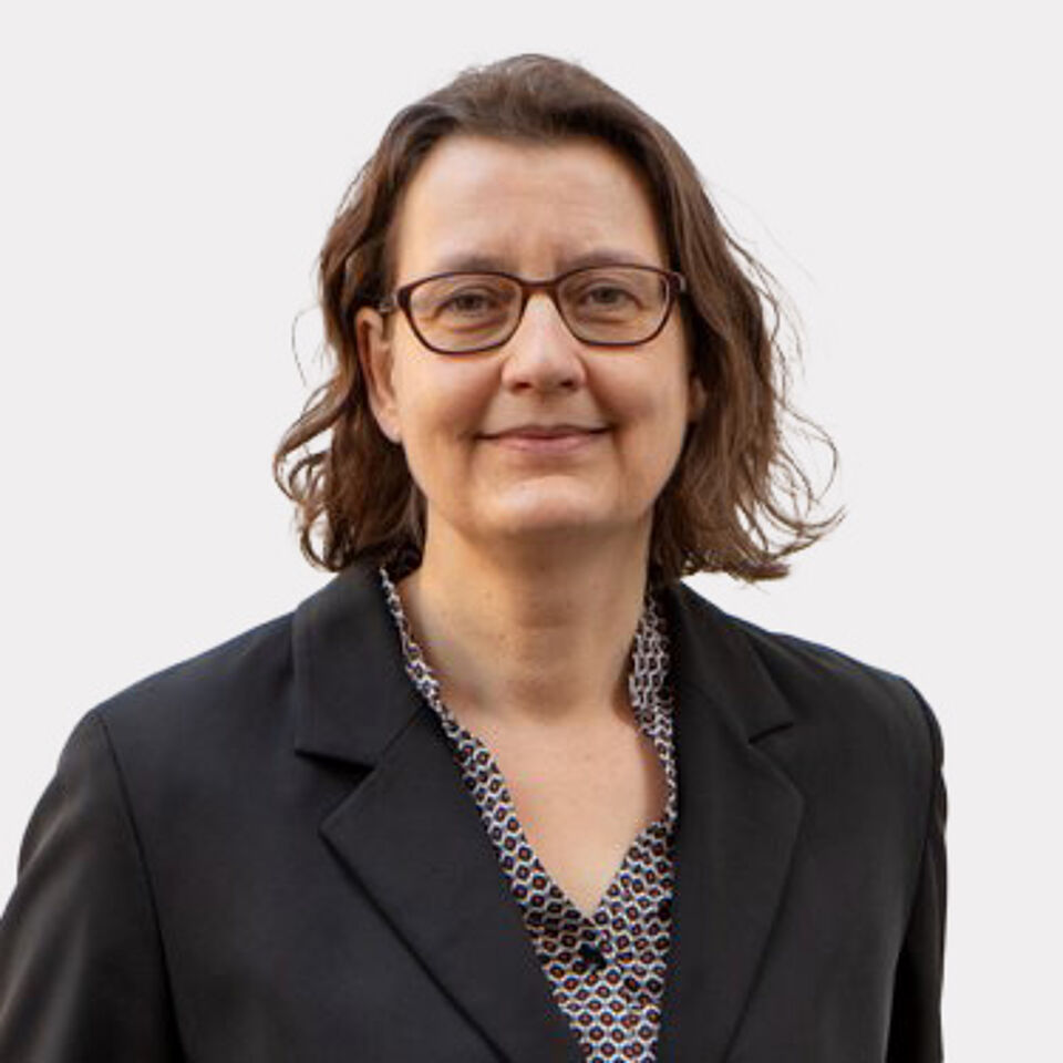 Frau mit braunen Haare und Brille mit gemusterter Bluse und schwarzem Blazer