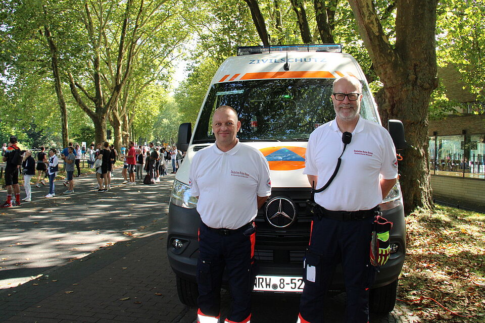 Marathon Zwei Personen in Einsatzkleidung vor einem Einsatzfahrzeug, im Hintergrund die Laufstrecke des Marathons
