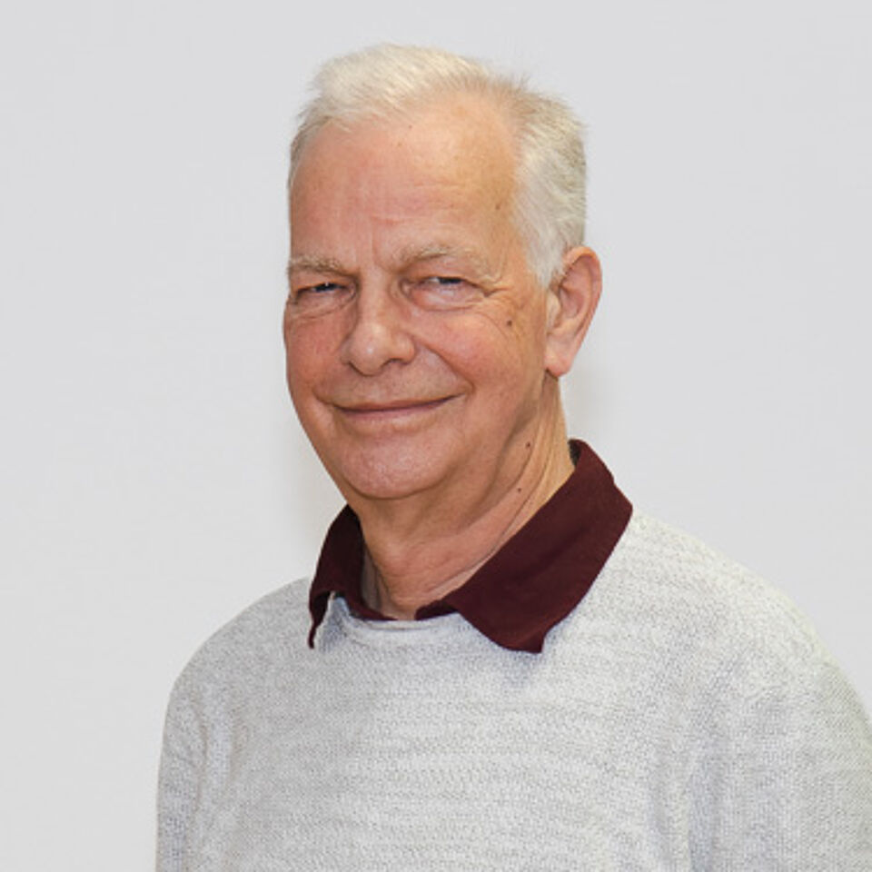 Mann mit weißen Haaren, einem grauen Pullover und rotem Hemdkragen vor weißem Hintergrund