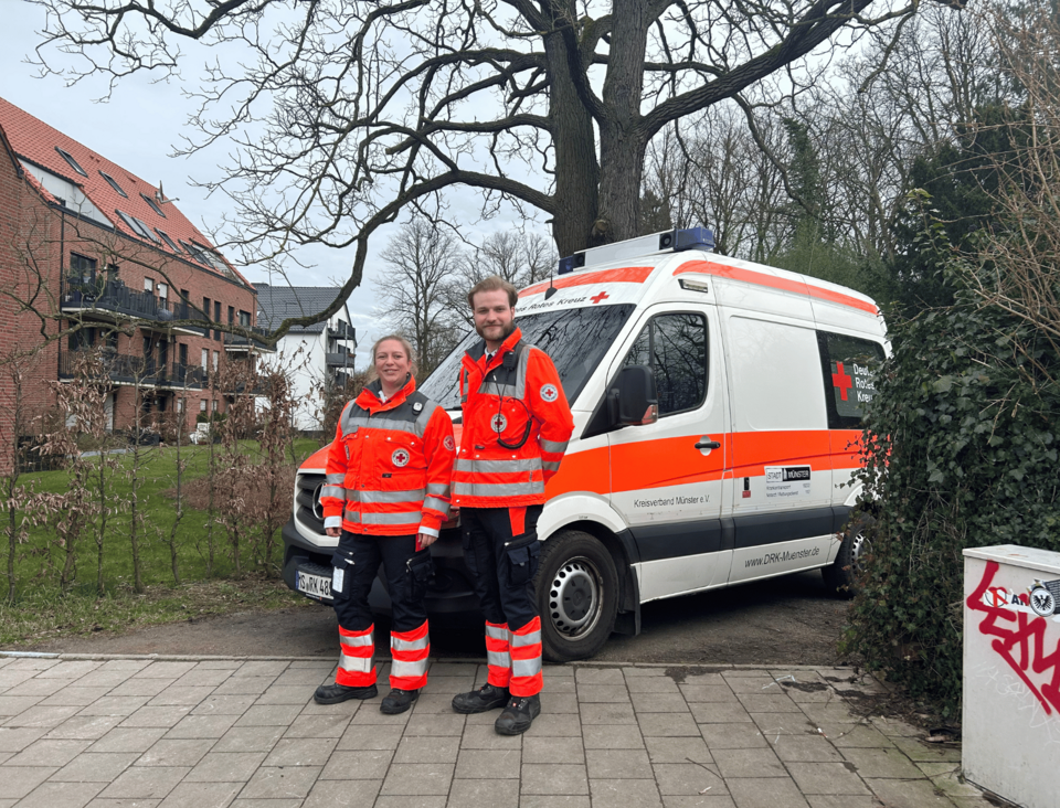 Zwei Einsatzkräfte vor einem Krankentransportwagen. Drumherum Bäume und Hecken.