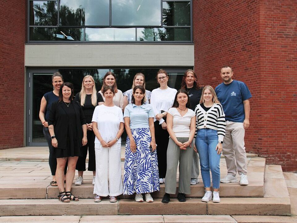Eine Gruppe von Personen auf Treppen vor einem Gebäude mit Fensterfront und Backsteinen.