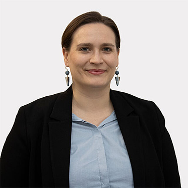 Frau mit braunen Haaren, blauer Bluse und schwarzem Blazer vor weißem Hintergrund