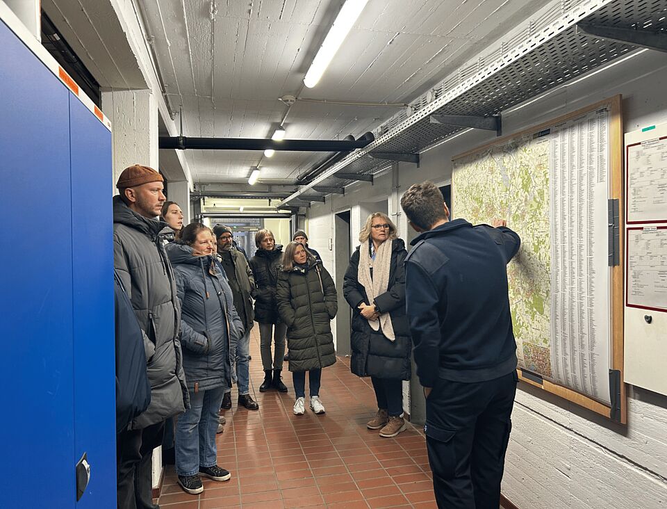 Eine Gruppe von Personen steht in einem Gang, an der Wand hängen Karten. Eine Person steht an den Karten und erklärt etwas.