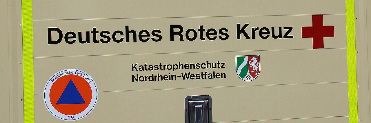 Ein Ausschnitt eines Fahrzeugs zeigt mehrere Aufschriften und Symbole. In der Mitte steht in schwarzer Schrift „Deutsches Rotes Kreuz“ neben einem roten Kreuz. Darunter befindet sich der Text „Katastrophenschutz Nordrhein-Westfalen“ sowie ein Wappen. Links ist ein rundes Emblem mit der Aufschrift „Medizinische Task Force 29“ zu sehen. In der Mitte dieses Emblems befindet sich ein blaues Dreieck in einem orangefarbenen Kreis.