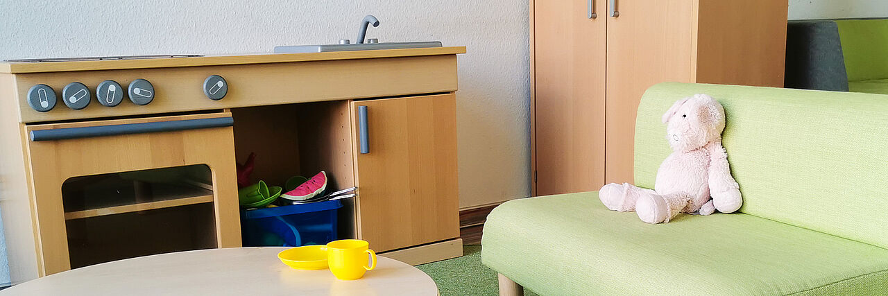 Erphobogen Slider Eine Spielküche, ein Sessel mit einem Kuscheltier, ein Schrank im Hintergrund