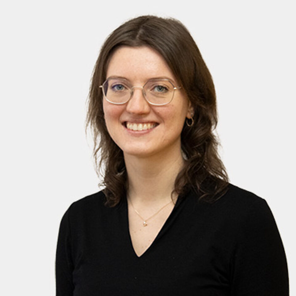 Frau mit braunen Haaren und Brille und schwarzem Oberteil vor weißem Hintergrund
