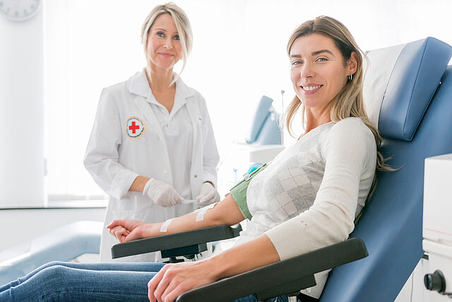 Zwei Personen befinden sich in einem medizinischen Umfeld. Eine der Personen trägt einen weißen Kittel mit einem Rotkreuz-Emblem auf der linken Seite, was auf eine Tätigkeit im Gesundheitswesen hinweist. Die andere Person sitzt und scheint medizinisch betreut oder beraten zu werden.