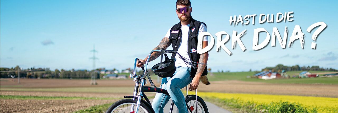 Eine Person fährt mit dem Fahrrad auf einer Landstraße umgeben von Feldern und einigen Gebäuden im Hintergrund. In den Speichen befinden sich Rotkreuzreflektoren. Text im Bild: „Hast du die DRK DNA?“
