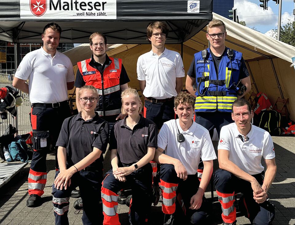 Acht Personen in zwei Reihen mit Einsatzkleidung vor einem Zelt mit der Aufschrift "Malteser, weil Nähe zählt"