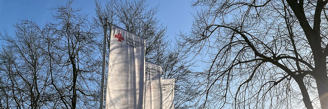 Drei weiße Fahnen mit DRK-Logo vor einem Baum und blauem Himmel.