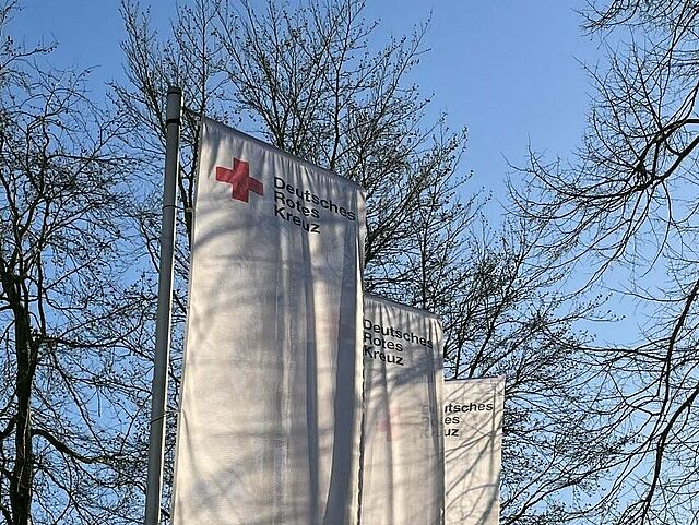 Drei weiße Fahnen mit DRK-Logo vor einem Baum und blauem Himmel.
