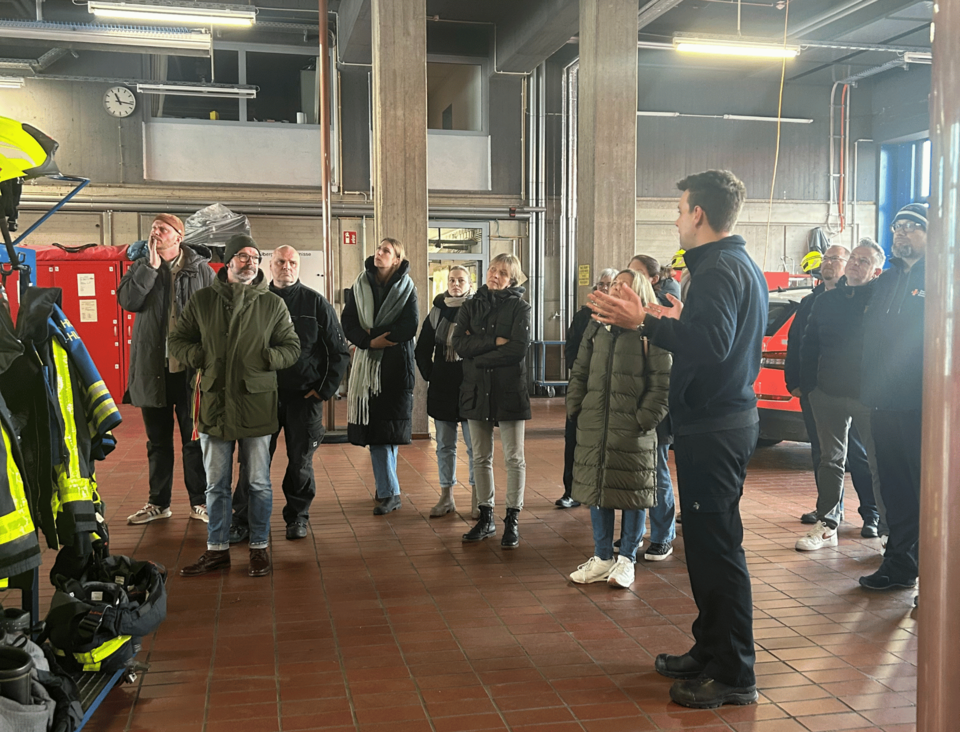 Eine Gruppe von Personen steht in der Halle einer Feuerwache. Eine Person steht vor der Gruppe und gibt eine Führung.