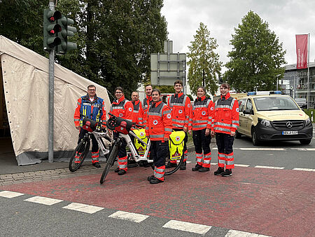 Gruppenbild von acht Einsatzkräften in Einsatzkleidung mit zwei Fahrrädern. Im Hintergrund sind ein Zelt und Autos zu sehen.