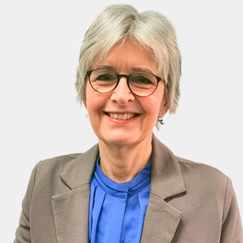 Frau mit weißen kurzen Haaren, Brille, blauer Bluse und grauem Blazer vor weißem Hintergrund