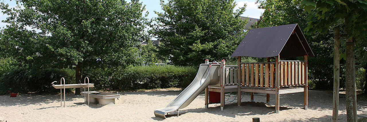 Meerwiese Spielplatz mit Sand, einem Häuschen mit Rutsche und Wasserspiel, im Hintergrund Bäume und Wohnbebauung