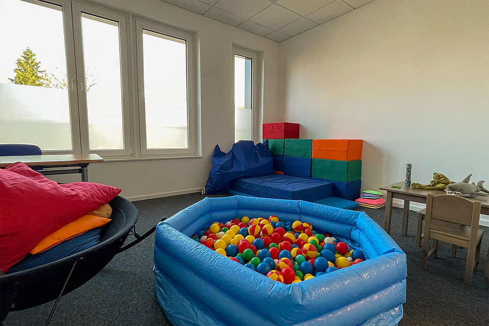 Ein Raum mit Fenster und einem bunten Bällebad mit blauer Umrandung, mehrere bunte Schaumstoffwürfel, ein Kindertisch mit 2 Stühlen.