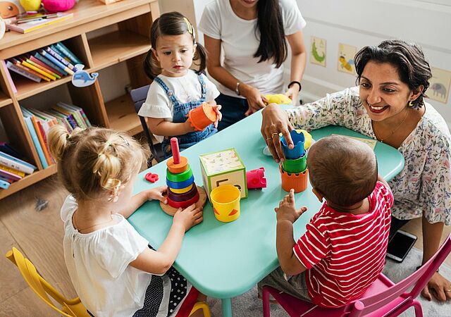 Mehrere Kinder und zwei Erwachsene sitzen um einen kleinen Tisch und beschäftigen sich mit buntem Spielzeug. Die Kinder spielen mit Stapelringen, Bauklötzen und Bechern. Im Hintergrund steht ein Regal, das mit Büchern und weiteren Gegenständen gefüllt ist.