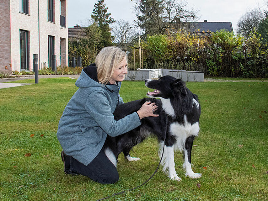 Eine Person kniet auf einer Wiese und macht einen Bodycheck bei einem schwarz-weißen Hund. Im Hintergrund sind ein modernes Gebäude, Bäume und Sträucher zu sehen.