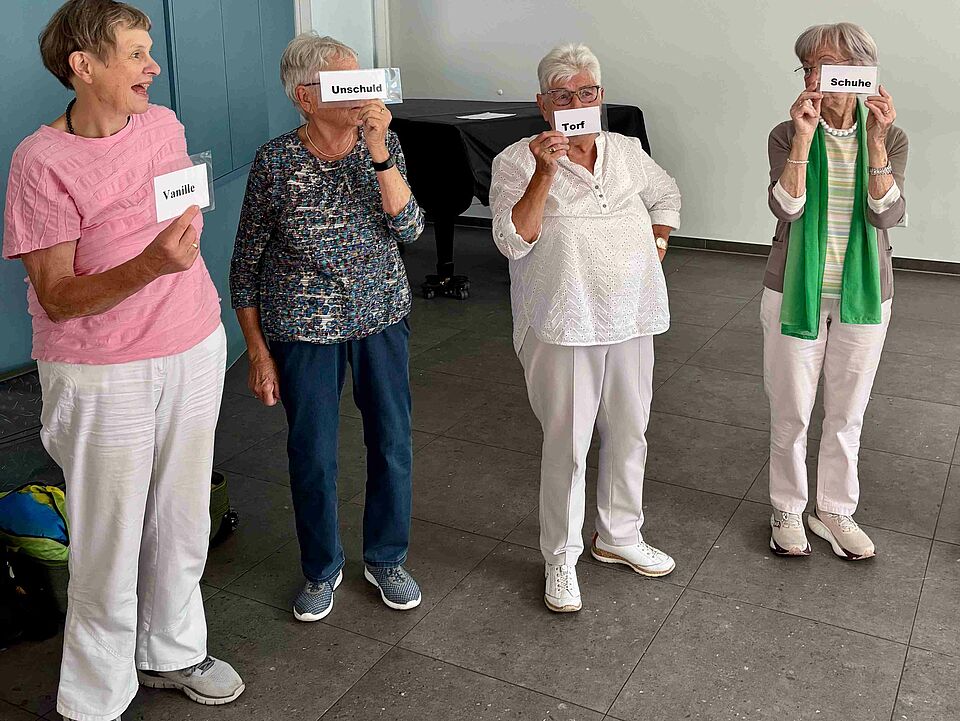 Vier älteren Frauen stehen in einer Reihe und halten Kärtchen mit Begriffen hoch
